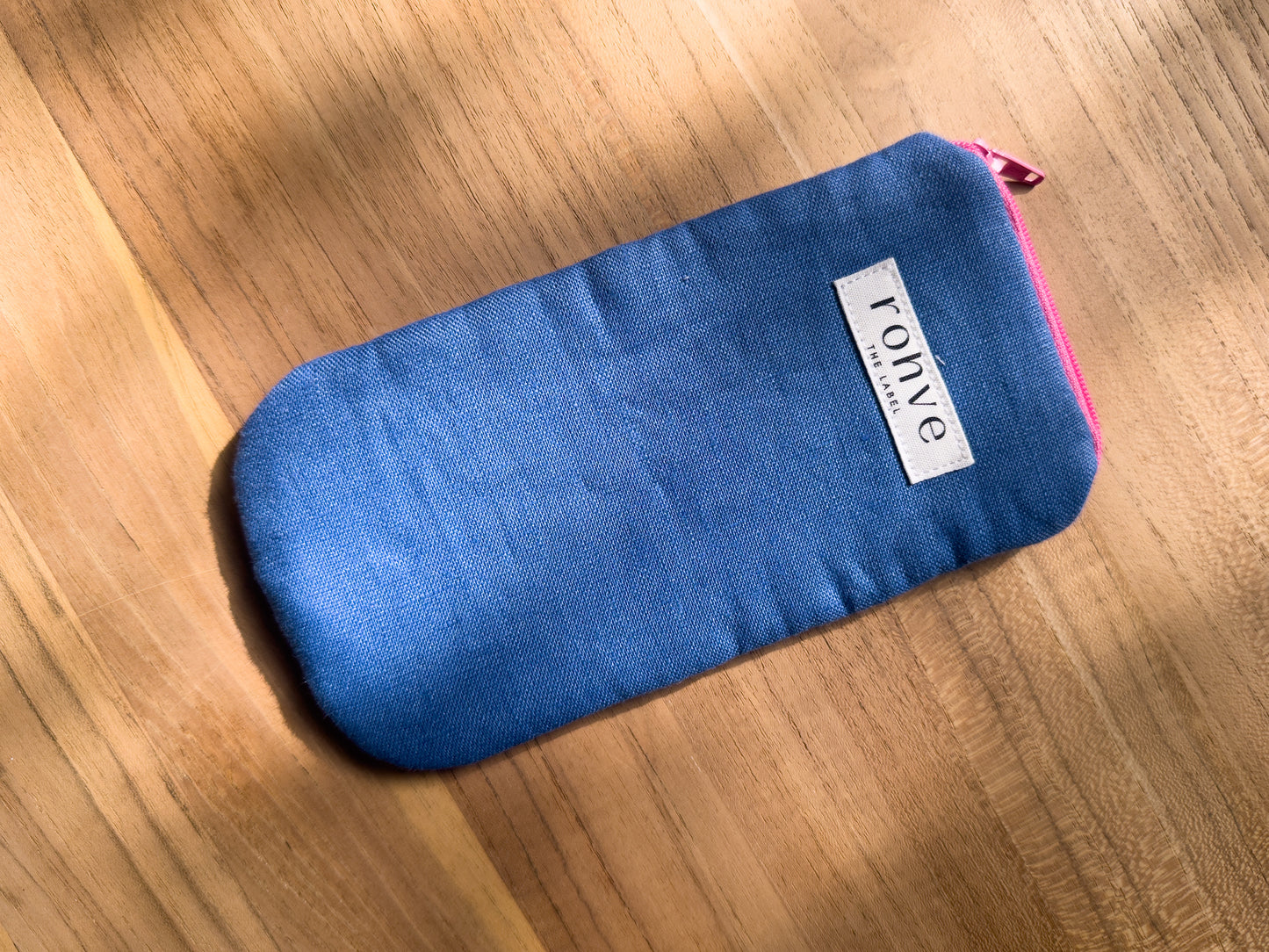 A mid blue linen fabric zippered pouch with a white brand label reading 'rohve' and a hot pink zipper, placed diagonally on a warm wooden surface.