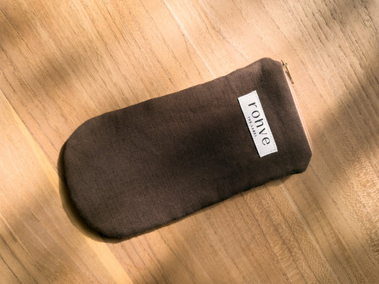 A brown linen fabric zippered pouch with a white brand label reading 'rohve' and a beige zipper, placed diagonally on a warm wooden surface.