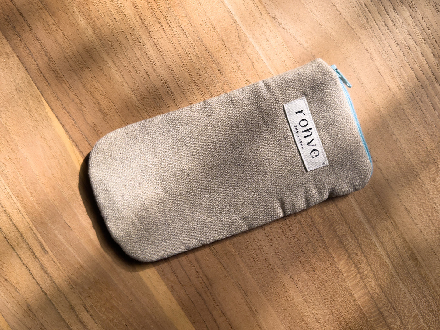 A beige linen fabric zippered pouch with a white brand label reading 'rohve' and a light blue zipper, placed diagonally on a warm wooden surface.
