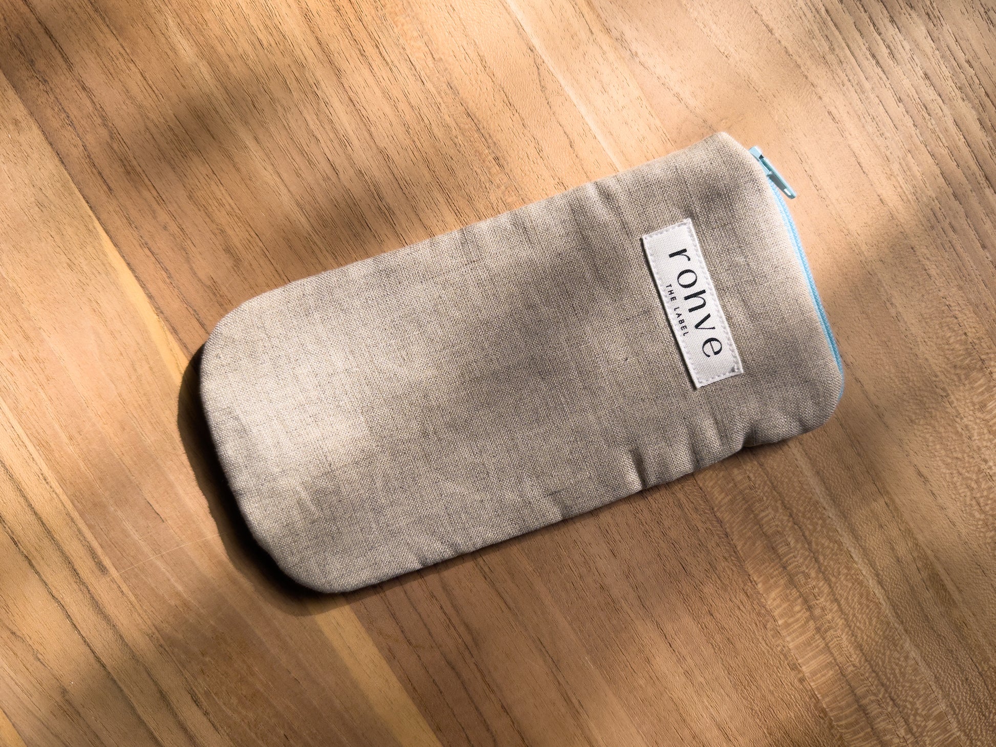 A beige linen fabric zippered pouch with a white brand label reading 'rohve' and a light blue zipper, placed diagonally on a warm wooden surface.