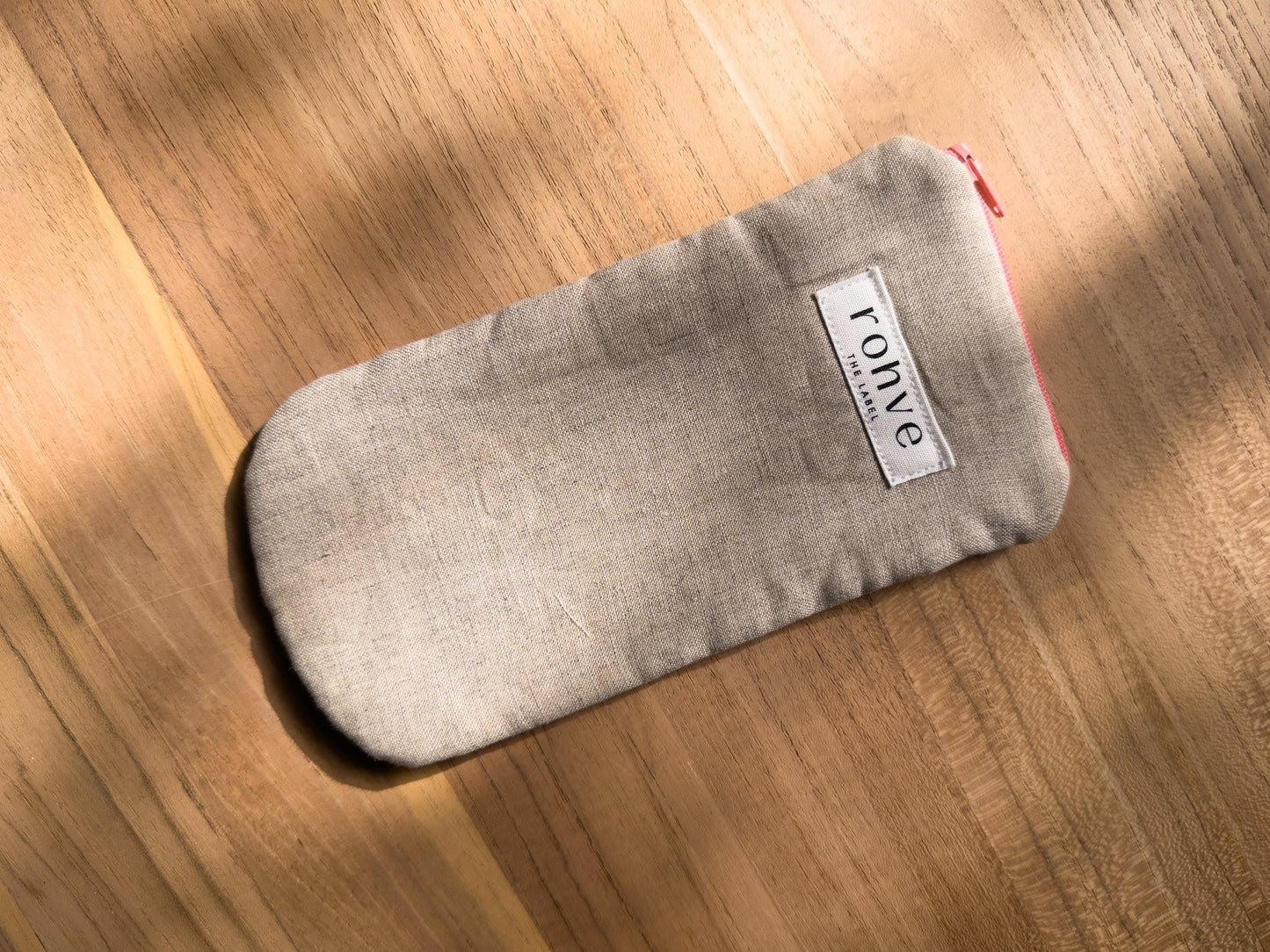 A beige linen fabric zippered pouch with a white brand label reading 'rohve' and a peach zipper, placed diagonally on a warm wooden surface.