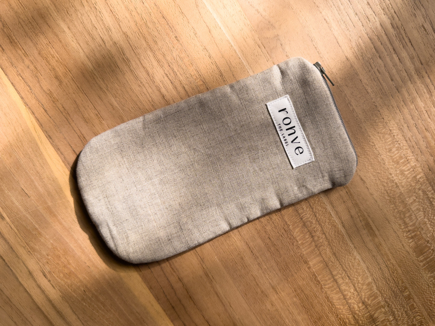 A beige linen fabric zippered pouch with a white brand label reading 'rohve' and a sage zipper, placed diagonally on a warm wooden surface.