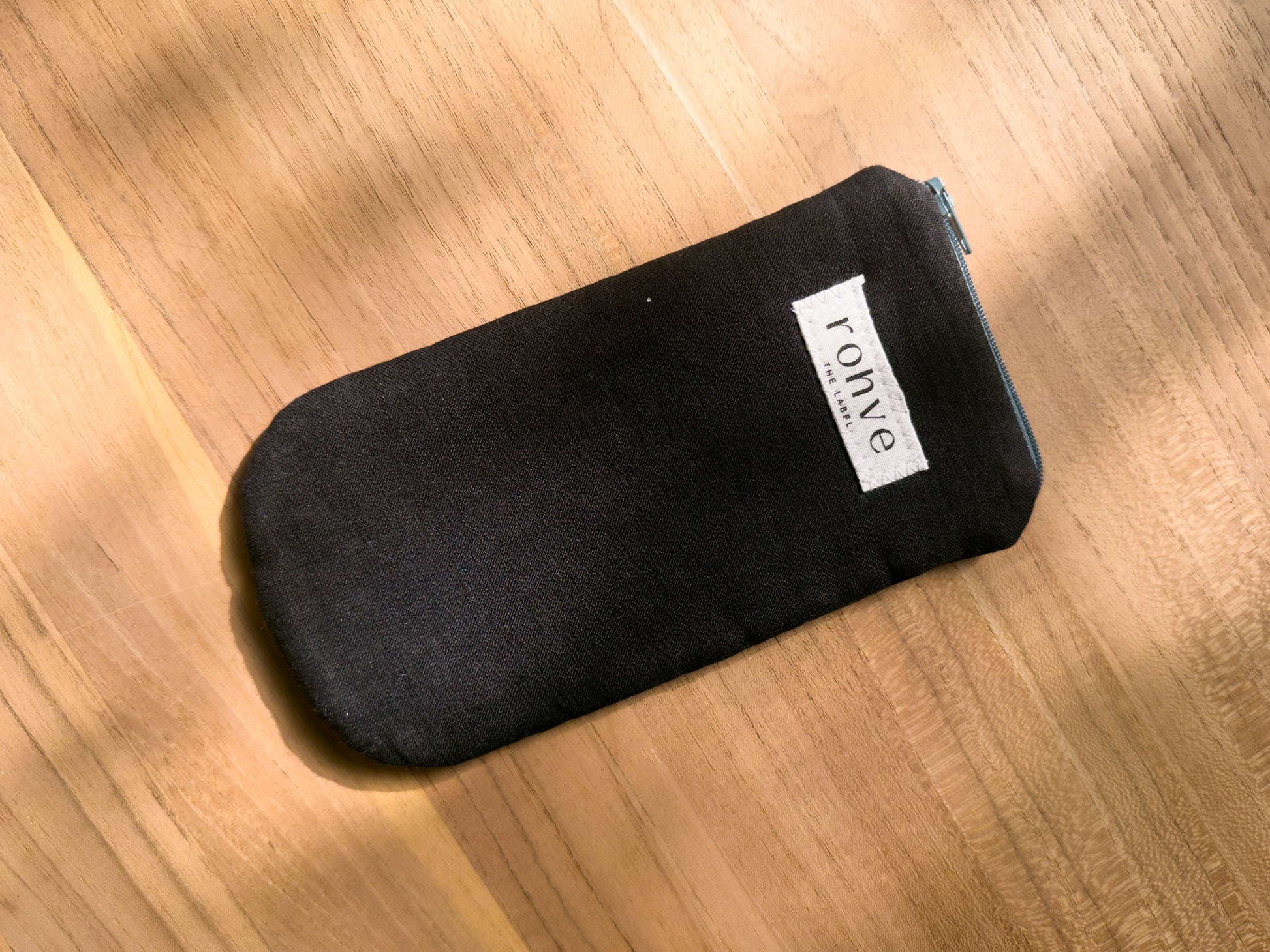 A black linen fabric zippered pouch with a white brand label reading 'rohve' and a teal zipper, placed diagonally on a warm wooden surface.