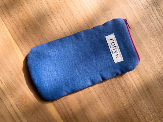 A mid blue linen fabric zippered pouch with a white brand label reading 'rohve' and a berry zipper, placed diagonally on a warm wooden surface.