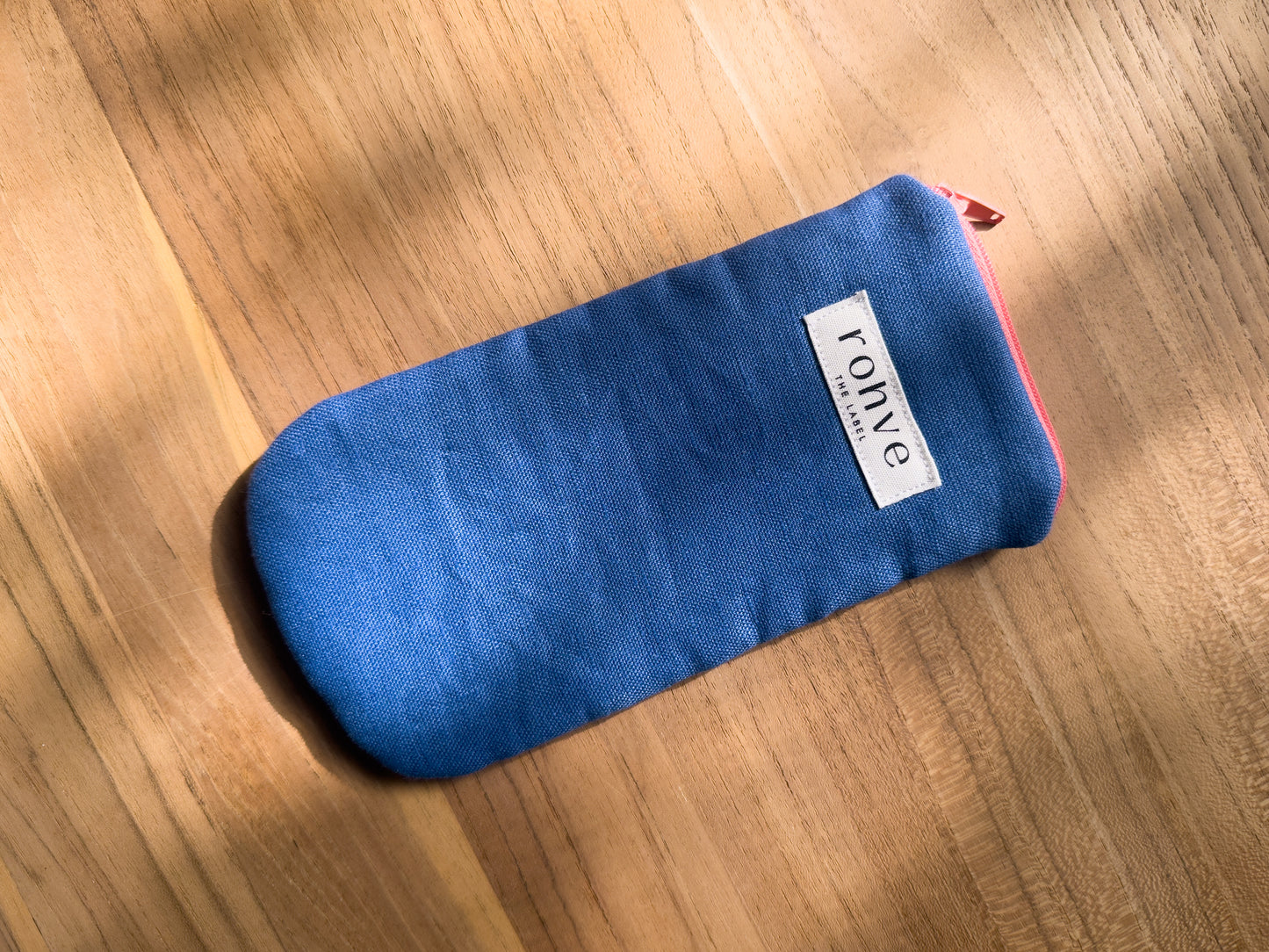 A mid blue linen fabric zippered pouch with a white brand label reading 'rohve' and a peach zipper, placed diagonally on a warm wooden surface.