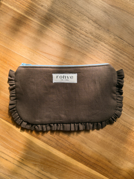 A brown fabric cosmetic pouch with ruffled trim, featuring a white label that reads 'rohve' on a wooden surface.