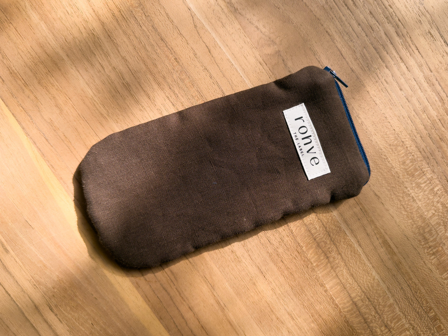A brown linen fabric zippered pouch with a white brand label reading 'rohve' and a navy zipper, placed diagonally on a warm wooden surface.